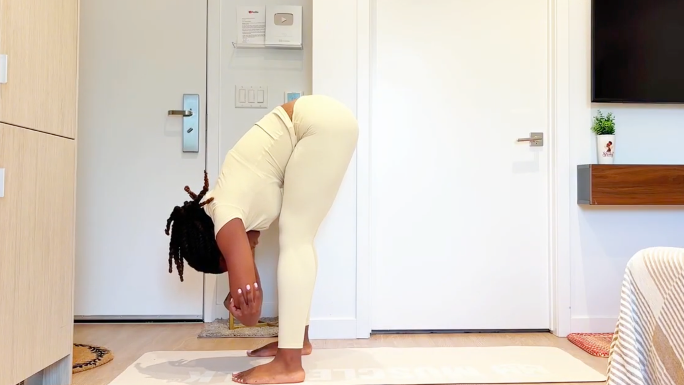 Yoga instructor Ramoni Overton doing Rag Doll pose in a standing forward bend while holding opposite elbows.